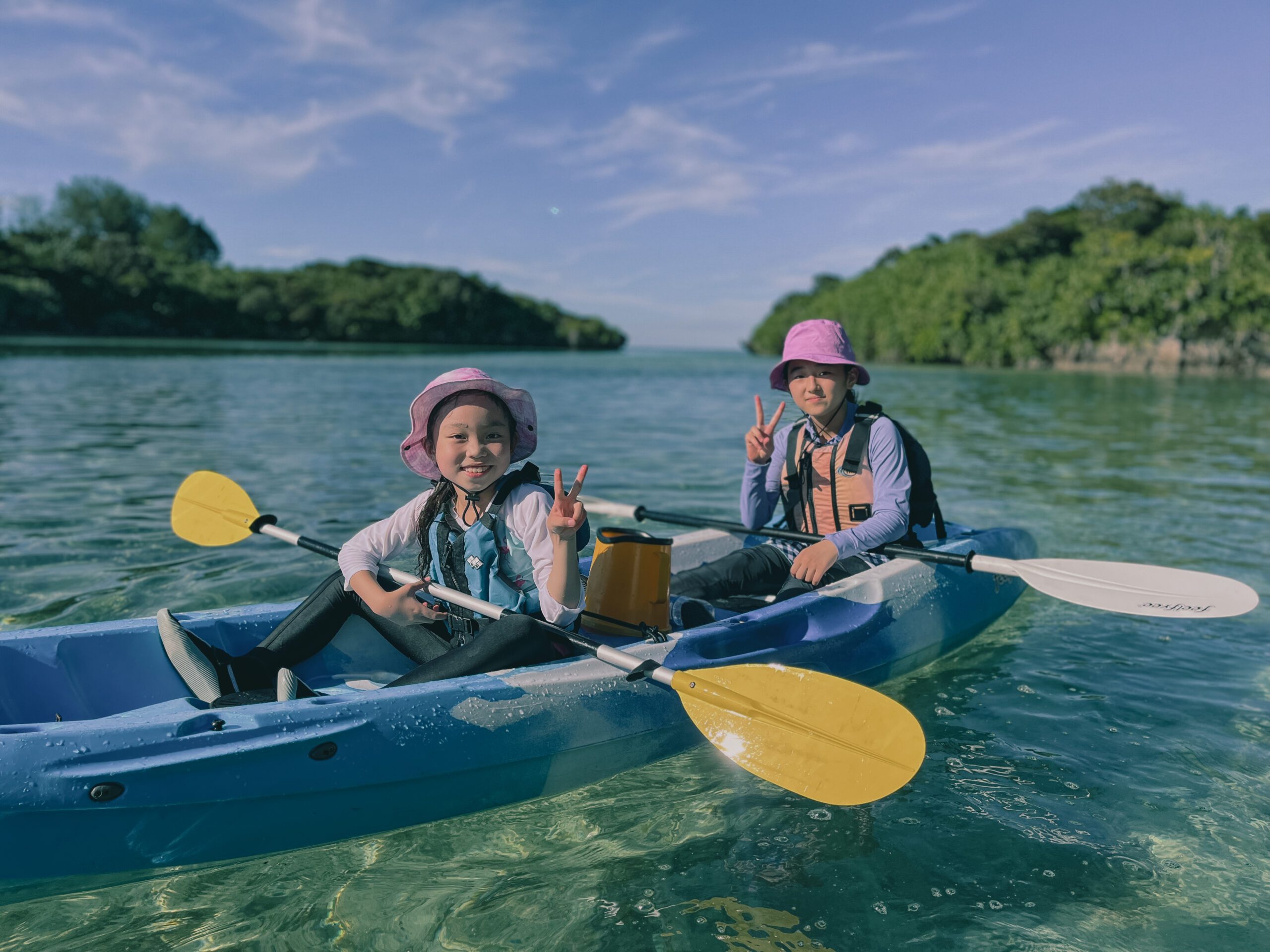 川平湾カヤックツーリングで透明な海に浮かぶカヤックからピースをする小学生の女の子二人。asobi基地ユニバーシティのプログラム「石垣島7日間生活」